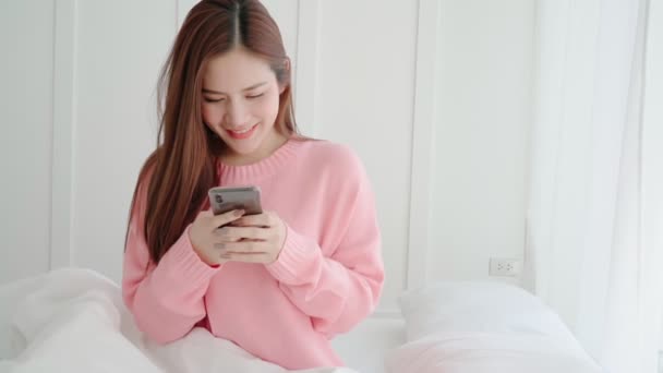 Красива довге волосся азіатська жінка приємна посмішка насолоджуйтесь чатом смартфона після пробудження ранкових вихідних на фоні інтер'єру спальні — стокове відео