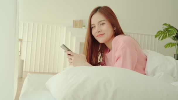 Bello lungo capelli asiatico donna bello sorriso godere smartphone chat dopo sveglia mattina fine settimana su letto camera da letto interno sfondo — Video Stock