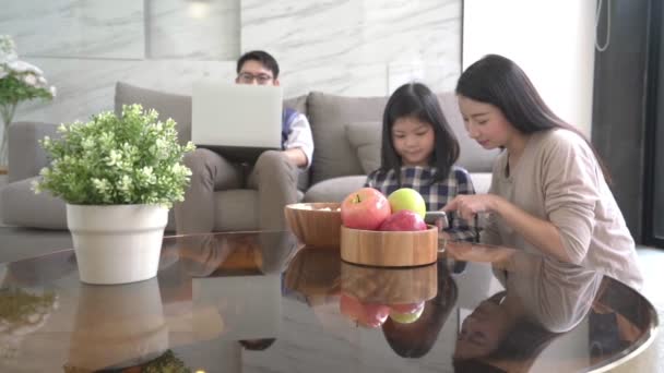 Повільний рух азіатська дочка робить домашнє завдання на блокноті. Його мати і батько сидять і дивляться на диван і використовують цифровий ноутбук разом на домашньому тлі . — стокове відео