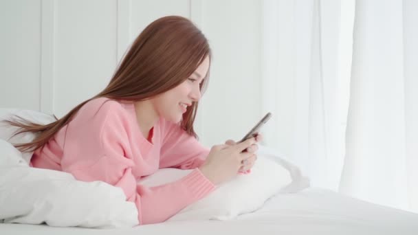 Όμορφη μακριά μαλλιά Ασιάτισσα γυναίκα ωραίο χαμόγελο απολαύσετε το smartphone chat μετά από ξύπνημα πρωί Σαββατοκύριακο στο κρεβάτι υπνοδωμάτιο εσωτερικό φόντο — Αρχείο Βίντεο