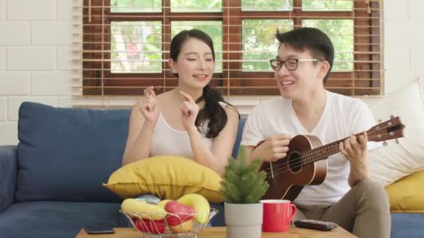 Молодой азиат жениться счастливая пара в игре на укулеле и петь в гостиной, счастье пара расслабляющий уик-энд вместе дома — стоковое видео