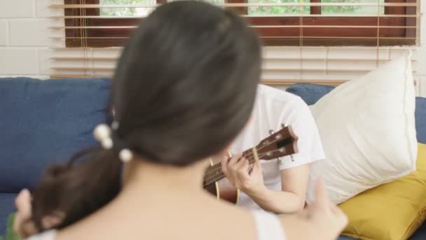 Młody Azji poślubić szczęśliwy para w gry ukulele i śpiewać w salonie, szczęście para relaksujący weekend czas razem w domu — Wideo stockowe
