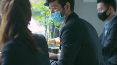 Yeni normal yaşam tarzı konsepti olan maske takan iş adamları sosyal mesafeli bir ortamda ya da bilgisayar ve akıllı telefonla ortak çalışma alanında oturuyorlar.