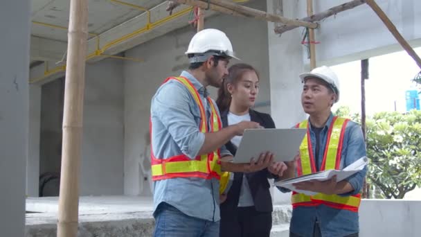 Giovani ingegneri asiatici e colleghi di ingegneri caucasici presso l'ispettore del sito e discutono insieme utilizzando il computer portatile e il progetto di carta presso il cantiere edilizio. — Video Stock