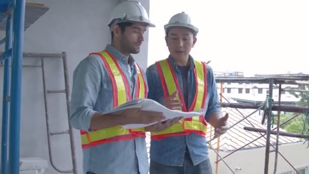 現地調査員の若手アジア人エンジニアと白人エンジニアの皆さんが、建築現場でノートパソコンと紙の青写真を使ってディスカッションを行います。ホームリノベーションのアイデアコンセプト — ストック動画