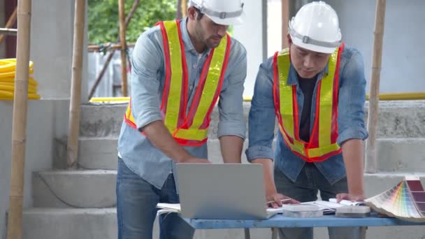 Young Asian engineers and caucasian engineer colleagues at site inspector and discuss together using notebook computer and paper blueprint at building construction site.home renovation ideas concept — Stock Video