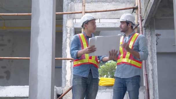 現地調査員の若手アジア人技術者と白人技術者が 建築現場でノートパソコンと紙の青写真を使って話し合います 住宅改修のアイデアコンセプト — ストック動画