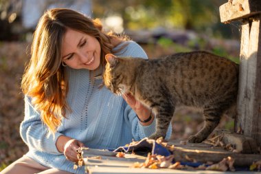 Açık mavi kazaklı, gülen genç bir kadın açık havada sıcak bir sonbahar gününün tadını çıkarıyor. Altın güneş ışığı ve sarı yapraklar altın bir düşüş ortamı yaratır..
