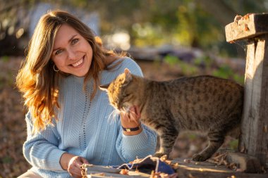 Açık mavi kazaklı gülümseyen genç bir kadının portresi güneşli bir sonbahar gününün tadını çıkarıyor. Altın yapraklar ve ılık güneş ışığı sonbahar mevsiminde yumuşak ve parlak bir atmosfer yaratır..
