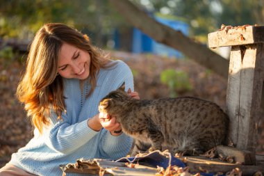 Açık mavi kazaklı, gülen genç bir kadın açık havada sıcak bir sonbahar gününün tadını çıkarıyor. Altın güneş ışığı ve sarı yapraklar altın bir düşüş ortamı yaratır..