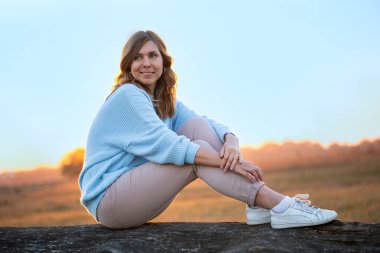 Açık mavi kazaklı genç bir kadının portresi güzel bir sonbahar günbatımında bir kütüğün üzerinde oturuyor. Sıcak altın ışık onun gülümsemesini ön plana çıkarıyor, sakin ve rahat bir açık hava ortamı yaratıyor..