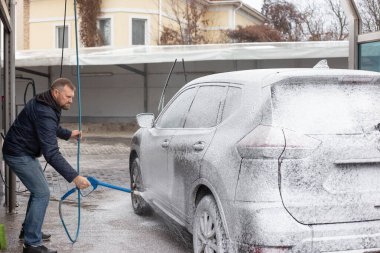 Otomobil yıkamada köpükle kaplı bir cipi temizleyen bir adam. Yüksek basınçlı hortumla soğuk bir günde aracı duruluyor. Açık hava ayarı, pratik oto bakım, etkin süreç.
