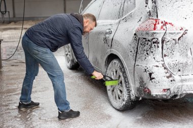 Adam oto yıkamada köpükle kaplı bir cipin tekerini fırçalıyor. Yeşil fırça kullanarak, ıslak günde kenarı iyice temizler. Pratik oto bakım, detay ve mevsimlik araç bakımı.
