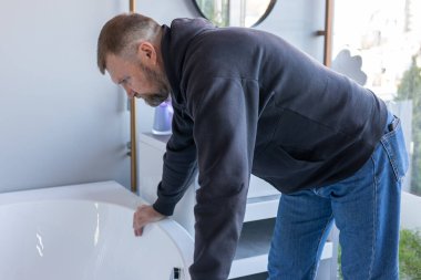 Su tesisatı dükkanında beyaz bir küveti inceleyen bir adam. Yeni bir küvet seçerken yüzeyi yakından incelemek için eğildi. Banyo armatürleri sergilenen parlak bir sergi salonu..