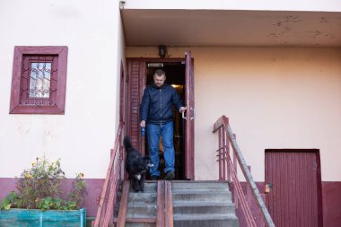 Bir adam köpeğiyle yürüyüşe çıkmak için apartmandan çıkıyor. Border collie melez köpek tasmayla önde yürüyor..