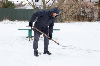 Soğuk kış gününde arka bahçeden kar küreyen orta yaşlı bir adam. Bir parka ve eldiven, mevsimlik bakım, kış işleri, ev bakımı, karlı bahçe işleri, dondurucu havada dışarıda işçilik..