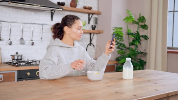 Morena saborear cereal usando celular na cozinha — Vídeo de Stock