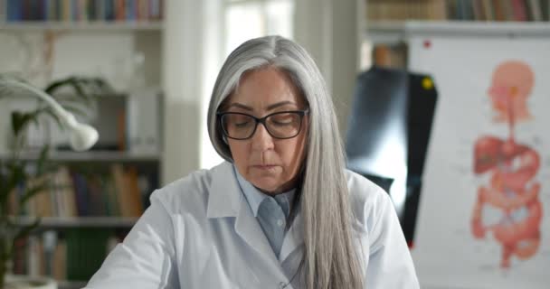 Close up of woman in white rob filling up medical form while having video online consultation. Elderly female doctor listening to patient and writing information while looking to camera. — Stock Video