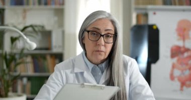 Hasta ile konuşan ve tıbbi form doldururken kameraya bakan yaşlı kadın doktor görüntüsü. Beyaz elbiseli kadın, elinde kağıt tutucu ve video ararken yazı yazıyor..