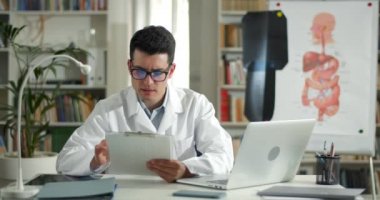 Beyaz elbiseli genç adam elinde kağıt tutuyordu ve online danışmanlık yaparken konuşuyordu. Erkek doktor görüntülü ararken konuşuyor ve tıbbi açıdan bakıyor. TeleHealth kavramı.