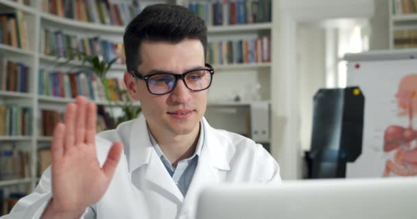 Gözlüklü bir adamın dizüstü bilgisayar ekranına bakarken el sallayıp konuşma görüntüsü. Tıp bürosunda online danışmanlık yaparken beyaz elbiseli neşeli erkek doktor yazı yazıyor.. — Stok video
