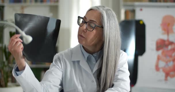 Crop view of female elderly doctor showing x ray photo while having online medical consultation. Mature lady in glasses looking to camera while communicating with patient in her office. — Stock Video