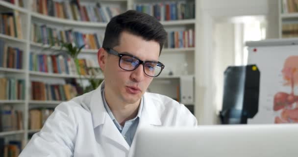 Cultivo hombre en gafas hablando mientras mira la pantalla del ordenador portátil. Hombre médico en vestido profesional blanco que tiene consulta en línea con el paciente en el consultorio médico. Concepto de telemedicina. — Vídeos de Stock