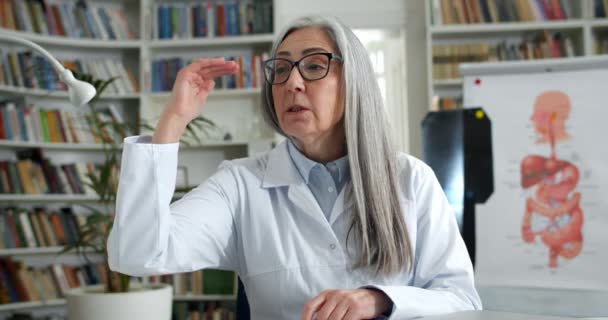 Close up view of elderly woman in glasses and white rob talking and gesturing while having online consultation. Female doctor looking to camera in medical office. Concept of telemedicine. — Stock Video