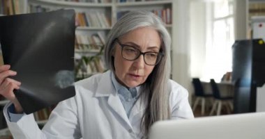 Ofiste tıbbi konsültasyon yapılırken kadın doktorun röntgen fotoğrafını gösterirken yakın plan. Gözlüklü ve beyaz önlüklü olgun bir kadın hastayla iletişim kurarken laptop kullanıyor..