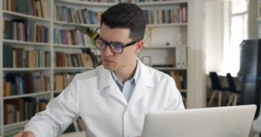 Erkek doktorun tıbbi belgelere baktığı ve ofiste otururken dizüstü bilgisayarda daktilo yazdığı görüntüleri kapatın. Gözlüklü genç adam ve iş yerindeki beyaz profesyonel soygunun görüntüsü.