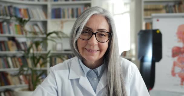 चिकित्सा कार्यालय में बैठे हुए कैमरा और मुस्कुराते हुए खुशबूदार बुजुर्ग महिला का चित्र। चश्मा और सफेद पेशेवर लूट में महिला डॉक्टर की फसल दृश्य। हेडशॉट की अवधारणा . — स्टॉक वीडियो