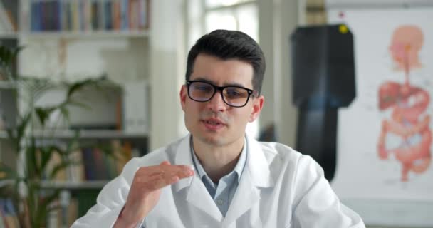 Close up view of young man in glasses and white rob talking and gesturing while having online consultation. Male doctor looking to camera in medical office. Concept of telemedicine. — Stock Video