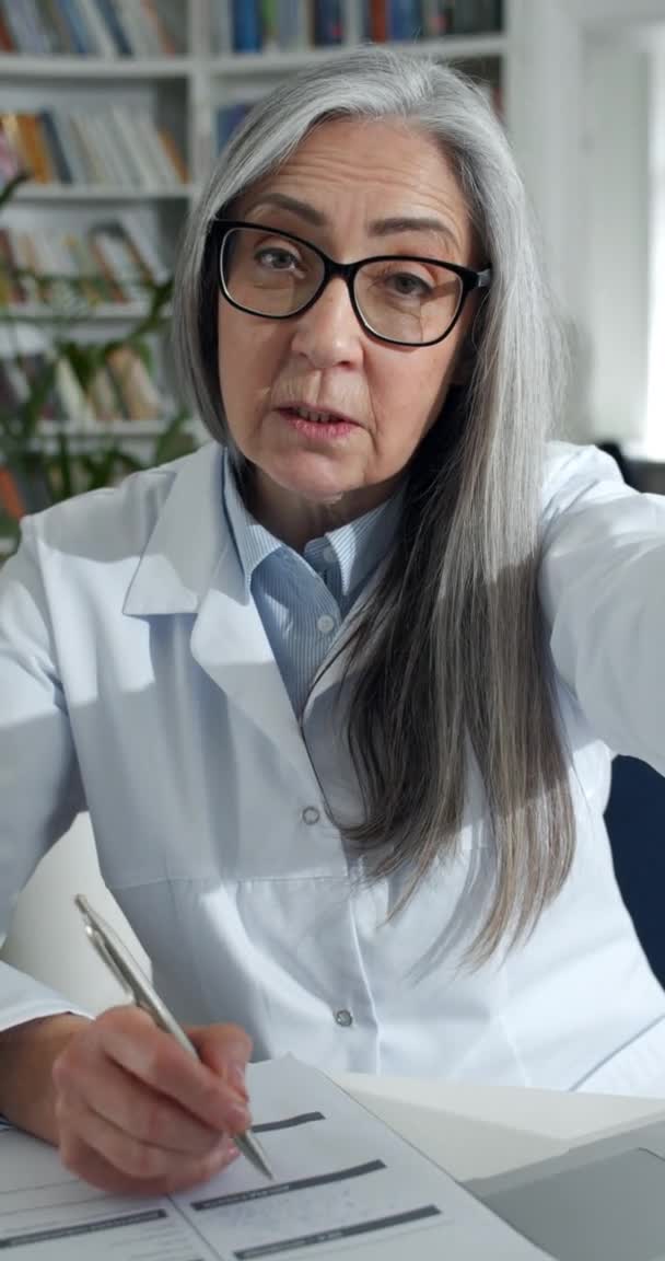Függőleges videó, amin idős nő szemüvegben és fehér köpenyben beszél és orvosi dokumentációt tölt ki. Női súlyos orvos dolgozik az irodában, miközben az online konzultáció. — Stock videók
