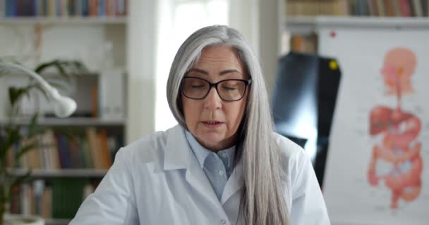 Crop view of professional woman in glasses and white gown gesturing and talking while having online conversation. Female doctor looking to camera in medical office. Concept of telehealth. — Stock Video