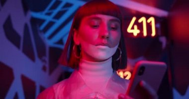 Akıllı telefon kullanan fütüristik makyajlı genç bir kadın. Burun halkalı bir kadın koridorda neon ışıkları ve parlayan numaralarla yürürken telefon ekranına bakıyor..
