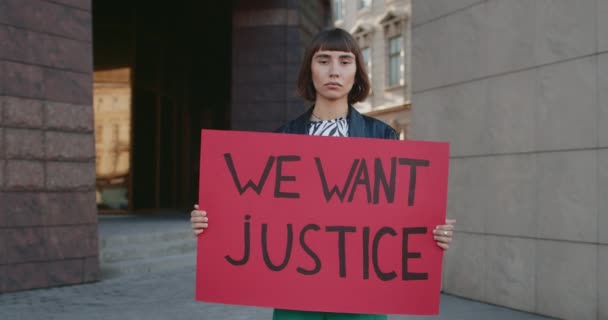 Retrato de una joven sosteniendo pancarta con nosotros queremos que la justicia escriba en ella. Hipster chica con anillo en la nariz que apoya el movimiento de derechos humanos mientras está de pie en la calle vacía de la ciudad. — Vídeos de Stock