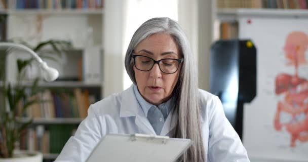 Crop view of woman in glasses taking and filling in patient medical form while having video call. Female doctor in white gown talking, writing and looking to camera in medical office. — Stock Video