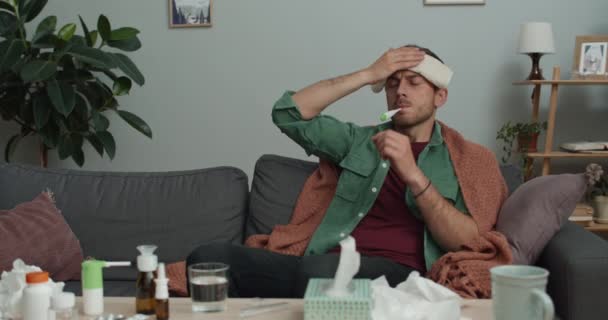 Nemocný chlap měřící tělesnou teplotu a držící tlak na čele, zatímco sedí doma na gauči. Vousatý muž podstupující léčbu při pohledu na termometr a třesoucí se hlavou. — Stock video