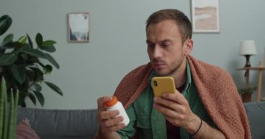 Soğuk sakallı adam battaniyeye sarılıp haplarla şişeye bakıyor ve gülümsüyor. 30 'lu yaşlarda hasta bir adam internet üzerinden ilaç kullanırken akıllı telefon kullanıyor..