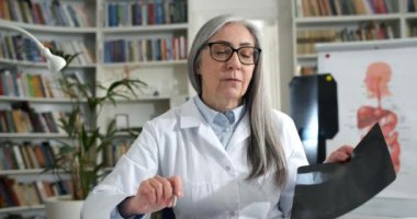 Doktorun gözlüklü görüntüsü hastaya röntgen fotoğrafını gösteriyor. Profesyonel beyaz önlüklü kadın kameraya bakıyor ve konuşuyor. Teletıp kavramı.