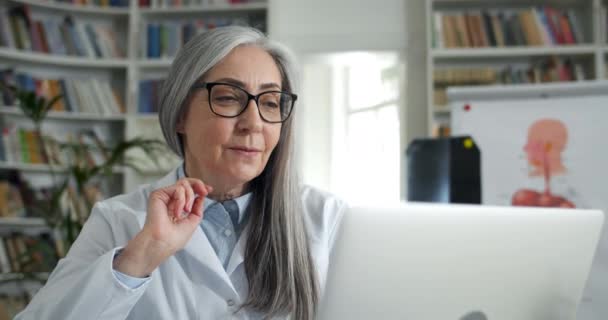 Crop view van volwassen vrouw in glazen zwaaien en praten tijdens het kijken naar laptop scherm. Volwassen vrouwelijke arts in witte jurk schrijven terwijl het hebben van online overleg in het medisch kantoor. — Stockvideo