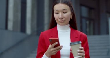 Akıllı telefon kullanan ve sokakta yürürken kağıt kahve fincanı taşıyan çekici bir iş kadını görüntüsü. Telefon ekranına dokunurken gülümseyen ve yan bakan Asyalı kadın..