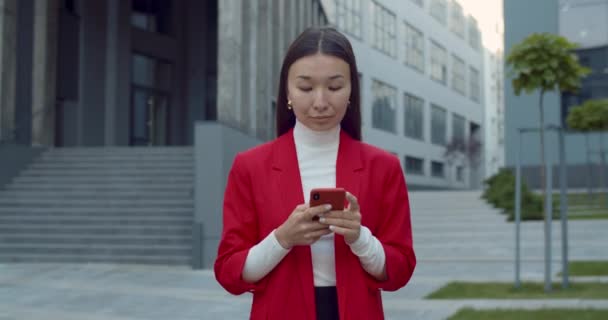 Vorderansicht einer hübschen asiatischen Geschäftsfrau, die ihr Smartphone auf der Straße hält und benutzt. Erfolgreiche Frau tippt und lächelt, während sie online kommuniziert und im Geschäftsbereich spaziert. — Stockvideo