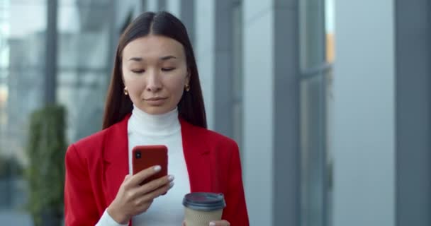 Primo piano di donna d'affari controllare il contenuto del telefono e guardando felice mentre si cammina per strada. Asiatico bella donna utilizzando smartphone e schermo di scorrimento durante il trasporto di caffè per andare. — Video Stock