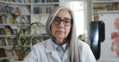 Tıbbi ofiste otururken kameraya bakan ciddi bir olgun kadının portresi. Kadın doktorun gözlüklü ve beyaz profesyonel önlüklü hali. Vesikalık fotoğraf kavramı.