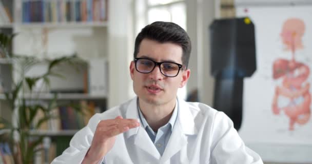 Serious young man in glasses and white professional coat talking and gesturing while having online consultation. Male doctor looking to camera in medical office. Telemedicine. — Stock Video