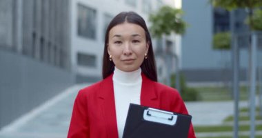 Asyalı güzel iş kadınının portresi. Elinde evrak klasörü var ve kameraya bakıyor. Ofis binalarının yakınındaki caddede duran başarılı kadın görüntüsü.