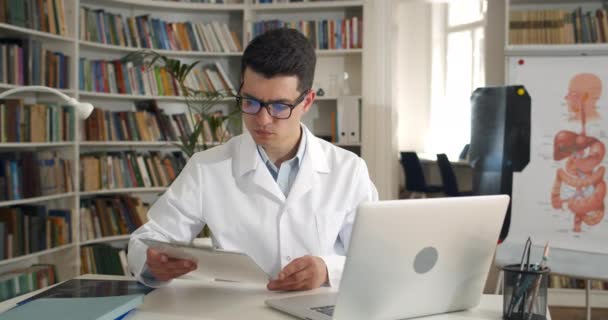 Tıbbi belgelere bakan ve ofiste otururken dizüstü bilgisayarda yazan bir erkek doktor. Gözlüklü genç bir adam ve iş yerinde beyaz profesyonel bir elbise. Tıp kavramı. — Stok video
