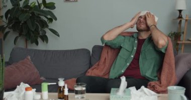 Soğuk algınlığı olan sakallı bir adam evdeki kanepede otururken kafasına kompres yapıyor. Battaniyeyle kaplanmış hasta evde tedavi görüyor. Sağlık ve hastalık kavramı.