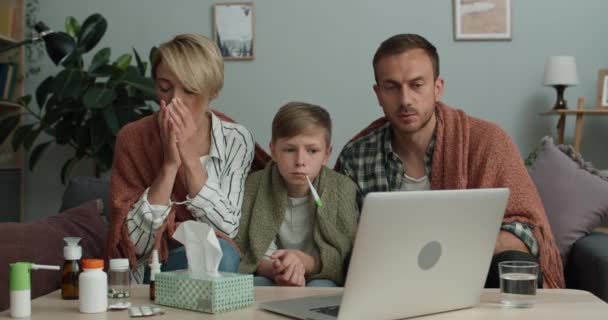 Madre, padre e figlio che hanno freddo e parlano durante la consultazione medica online. Famiglia malata in cerca di fotocamera del computer portatile mentre seduto sul divano a casa. Concetto di telemedicina. — Video Stock